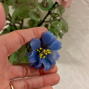 May include: A close-up of a blue flower with yellow stamens, held in a hand. The flower is part of a larger arrangement with green leaves and pink flowers. The background is a white, textured surface.