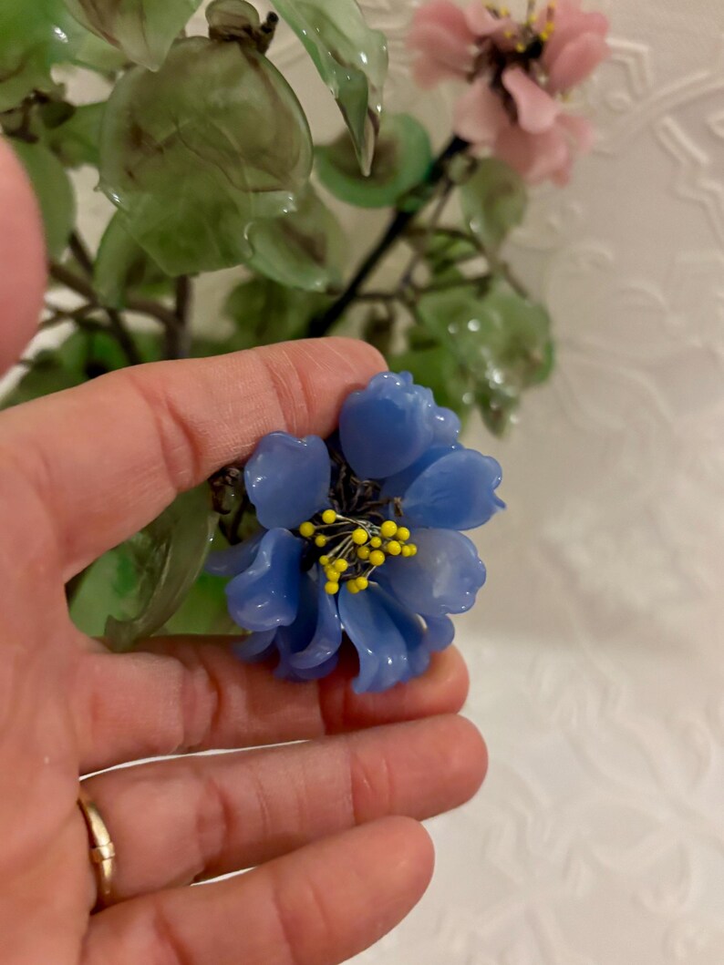 May include: A close-up of a blue flower with yellow stamens, held in a hand. The flower is part of a larger arrangement with green leaves and pink flowers. The background is a white, textured surface.