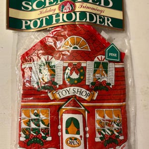 May include: A red and white holiday pot holder shaped like a toy shop. The pot holder has a Christmas theme with images of wreaths, bows, and Christmas trees. The packaging reads "Scented Holiday Trimmings Pot Holder".