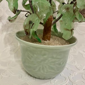 May include: A decorative bonsai tree in a light green ceramic pot. The tree features translucent green leaves and a brown trunk. The pot has a textured floral design and is filled with sand. The overall aesthetic is a blend of nature and artistry.