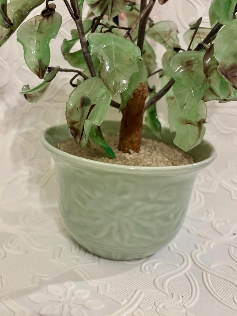 May include: A decorative bonsai tree in a light green ceramic pot. The tree features translucent green leaves and a brown trunk. The pot has a textured floral design and is filled with sand. The overall aesthetic is a blend of nature and artistry.