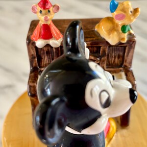 May include: A ceramic figurine featuring Mickey Mouse in a tuxedo, with a piano and two other cartoon characters. The figurine has a glossy finish with bright colors, including black, white, red, and yellow. The piano is brown with white keys.