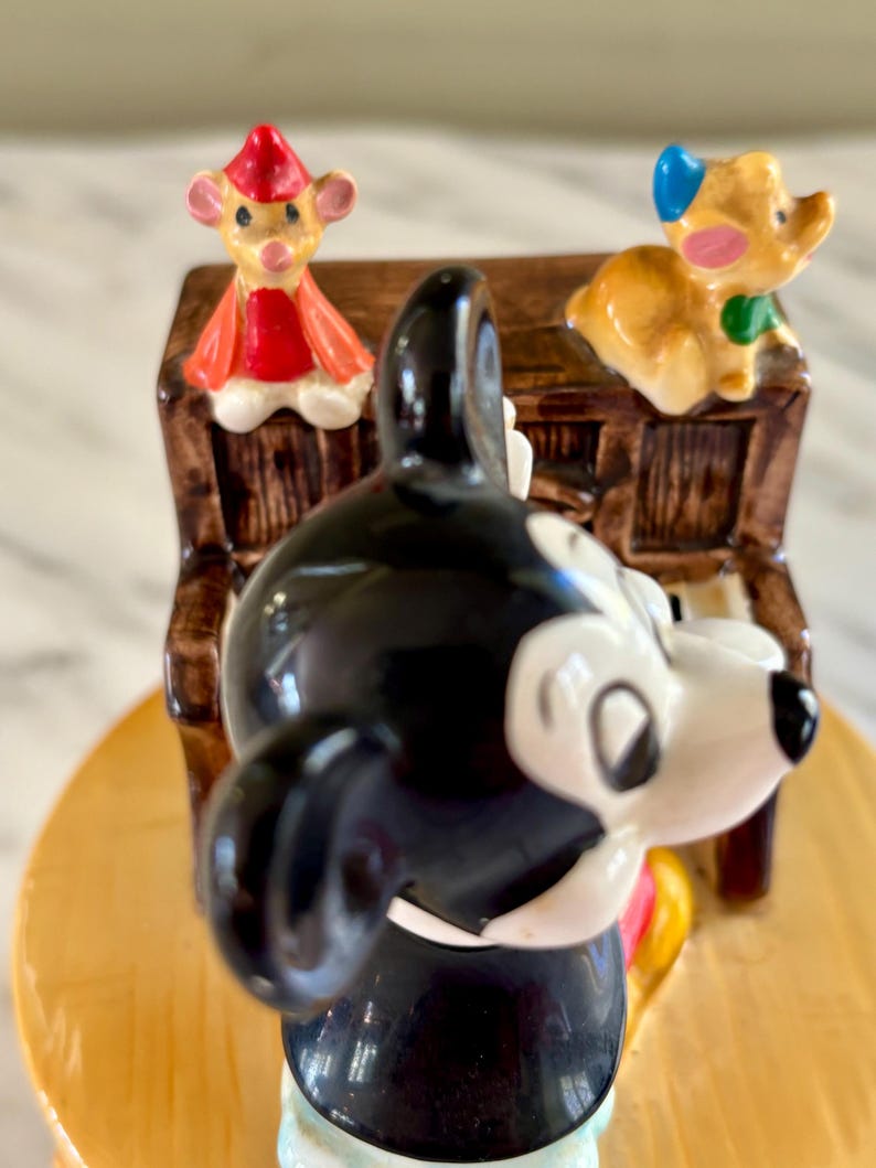 May include: A ceramic figurine featuring Mickey Mouse in a tuxedo, with a piano and two other cartoon characters. The figurine has a glossy finish with bright colors, including black, white, red, and yellow. The piano is brown with white keys.