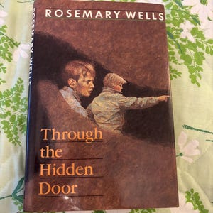 May include: A hardcover book titled "Through the Hidden Door" by Rosemary Wells. The cover features an illustration of two boys in a brown and tan color scheme. The book is set against a floral background.