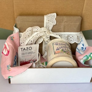 May include: A white gift box with a pink and green fabric on top. The box contains a white ceramic mug with the words "Great Adventure" printed on it and a box of Tazo Regenerative Organic Awake English Breakfast tea.