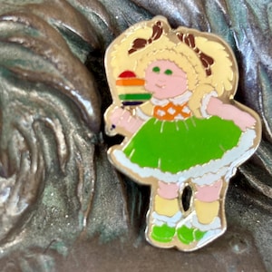 May include: A gold-tone pin featuring a cartoon girl with blonde hair, green eyes, and a pink dress. She is holding a rainbow lollipop.