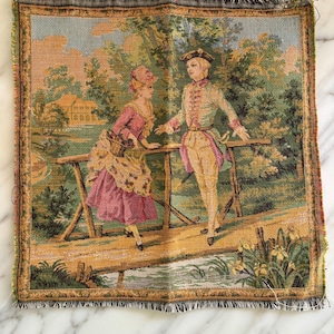 May include: A vintage tapestry depicting a couple in 18th-century clothing standing on a wooden bridge over a stream. The woman is wearing a pink dress and a white hat, while the man is wearing a brown and white suit. The background features a lush green forest and a small house.