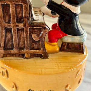 May include: A ceramic figurine of Mickey Mouse playing a piano. Mickey is wearing a black jacket, red shorts, yellow shoes, and white gloves. The piano is brown with white keys. The base is yellow with musical notes and the text "WALT DISNEY PRODUCTIONS. JAPAN".