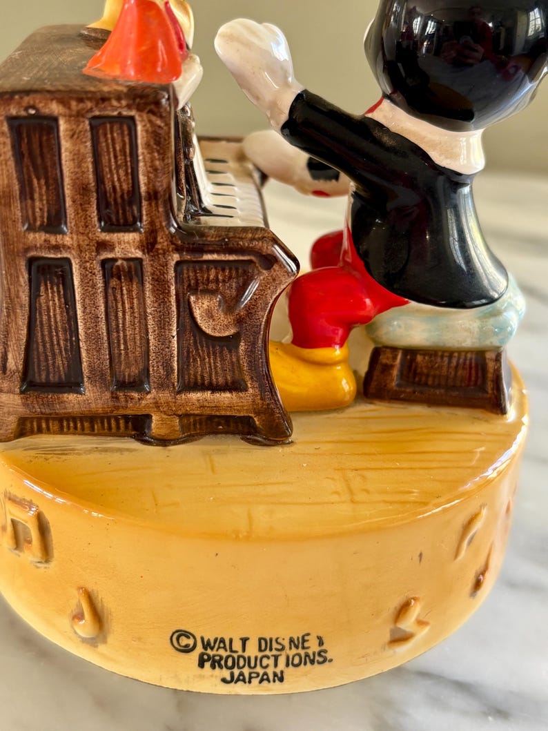 May include: A ceramic figurine of Mickey Mouse playing a piano. Mickey is wearing a black jacket, red shorts, yellow shoes, and white gloves. The piano is brown with white keys. The base is yellow with musical notes and the text "WALT DISNEY PRODUCTIONS. JAPAN".
