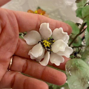 May include: A close-up of a white flower with yellow and black accents in the center. The flower has multiple petals and is part of a larger floral arrangement. Green leaves are also visible.