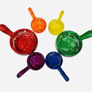 May include: A collection of six glitter-filled scoops in a rainbow of colors: red, orange, yellow, green, blue, and purple. Each scoop has a small handle and a round bowl shape, perfect for measuring ingredients or decorative use.