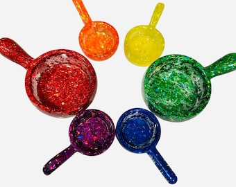 May include: A collection of six glitter-filled scoops in a rainbow of colors: red, orange, yellow, green, blue, and purple. Each scoop has a small handle and a round bowl shape, perfect for measuring ingredients or decorative use.