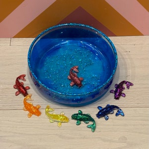May include: A blue glass bowl with a glittery surface, filled with water and surrounded by six colorful fish figurines. The fish are red, orange, yellow, green, purple, and blue.