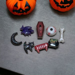 May include: A collection of Halloween-themed resin charms, including a black cat, a coffin, a cauldron, a crescent moon, a bat, an eyeball, and a fang. The charms are decorated with glitter and other embellishments.