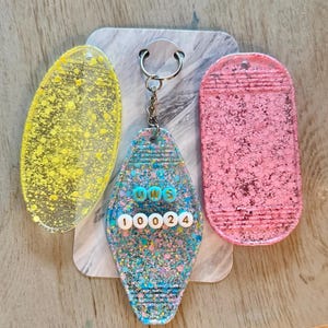 May include: A collection of three keychains. One is yellow with glitter, another is pink with glitter, and the third is a motel-style keychain with the text "UWS 10024" in colorful beads. The keychains are on a marble-patterned background.
