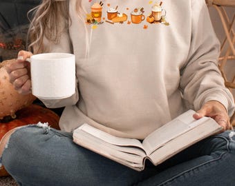 May include: A woman wearing a beige sweatshirt with a graphic of fall-themed drinks and pumpkins. She is sitting on a couch with a book in her lap.