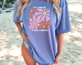 May include: A light purple t-shirt with a graphic of pink and orange flowers and the text "It's a good day to have a good day."