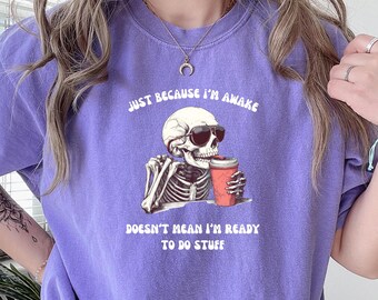 May include: Purple t-shirt featuring a skeleton wearing sunglasses and holding a cup of coffee. The text on the shirt reads "Just because I'm awake doesn't mean I'm ready to do stuff."