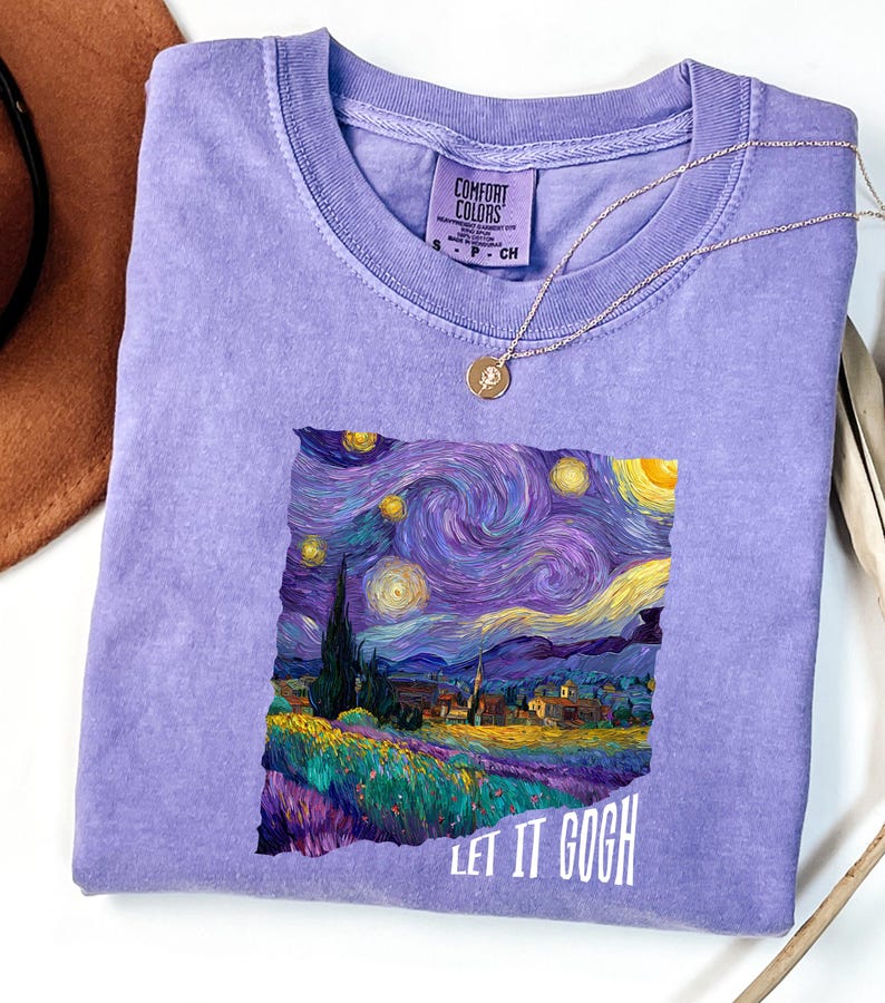 K&ouml;nnte beinhalten: Ein fliederfarbenes Comfort Colors T-Shirt mit einem Druck von Van Goghs "Sternennacht"-Gem&auml;lde. Das Kunstwerk zeigt wirbelnde Blau-, Lila- und Gelbt&ouml;ne mit dem Text "LET IT GOGH" in Wei&szlig;. Eine goldene Halskette liegt auf dem Shirt.
