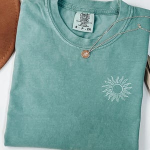 May include: A teal blue t-shirt with a white sun graphic. The shirt has a Comfort Colors tag with the size S-P-CH.