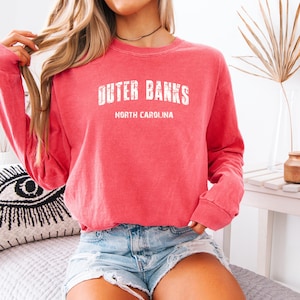 May include: A coral-colored long-sleeve sweatshirt with the text "OUTER BANKS NORTH CAROLINA" in white. The sweatshirt is worn with denim shorts. The image is taken indoors.