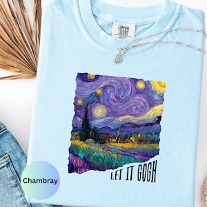 K&ouml;nnte beinhalten: Hellblaues Comfort Colors T-Shirt mit einem lebendigen Druck von Van Goghs Gem&auml;lde "Sternennacht". Das Design zeigt wirbelnde Blau-, Lila- und Gelbt&ouml;ne mit dem Text "LET IT GOGH" in Schwarz. Eine silberne Halskette mit Sonnenanh&auml;nger ist sichtbar.