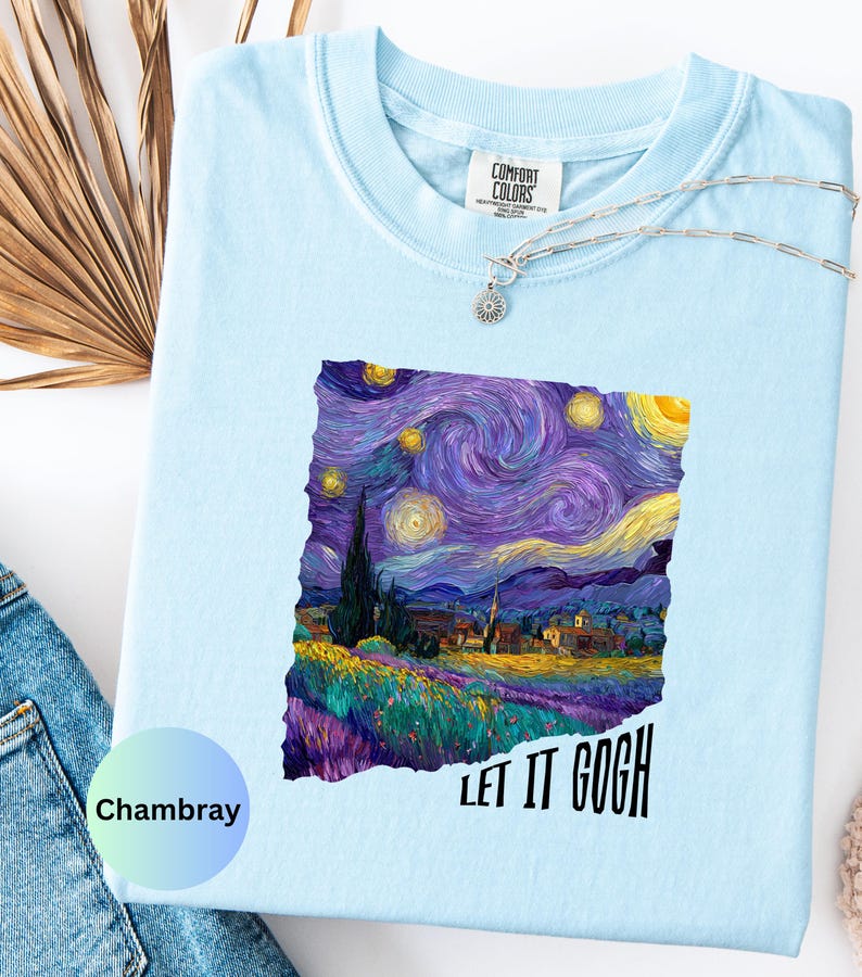 K&ouml;nnte beinhalten: Hellblaues Comfort Colors T-Shirt mit einem lebendigen Druck von Van Goghs Gem&auml;lde "Sternennacht". Das Design zeigt wirbelnde Blau-, Lila- und Gelbt&ouml;ne mit dem Text "LET IT GOGH" in Schwarz. Eine silberne Halskette mit Sonnenanh&auml;nger ist sichtbar.
