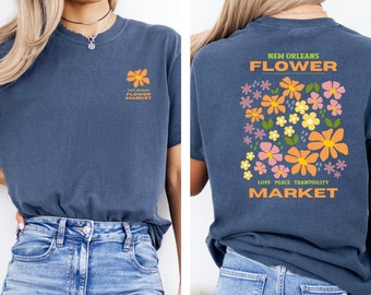 Puede incluir: Una camiseta azul marino con un diseño floral en la espalda. El diseño presenta una variedad de flores en tonos naranja, rosa, amarillo y verde. El texto "NEW ORLEANS FLOWER MARKET" está impreso en la parte posterior de la camiseta. El texto "LOVE PEACE TRANQUILITY" está impreso debajo de las flores.