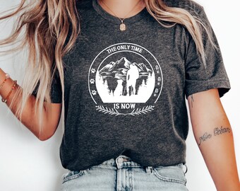 May include: A gray t-shirt with a white graphic design of a person and a dog hiking in the mountains. The graphic is in a circle with the text "The Only Time Is Now" and paw prints around the circle.