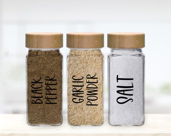 May include: Three glass spice jars with bamboo lids, labelled "BLACK PEPPER," "GARLIC POWDER," and "SALT." The jars contain dark brown, tan, and white ingredients. The labels are in a simple, black font.