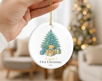 May include: A round white ceramic ornament featuring a watercolor illustration of a teddy bear sitting on gift boxes in front of a blue-lit Christmas tree. The text reads Liam's First Christmas 2026 in black and gold script and print.
