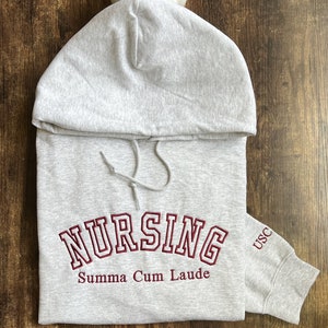 Peut inclure: Sweat-shirt à capuche gris avec les mots "Nursing Summa Cum Laude" brodés en bordeaux sur le devant et "USC" brodés en bordeaux sur la manche.