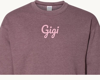 May include: A heathered maroon crewneck sweatshirt featuring the word Gigi embroidered in light pink script lettering across the center chest. The garment has a ribbed collar and long sleeves with a visible brand label at the neckline.