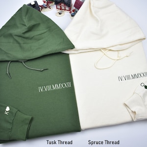 May include: Two hooded sweatshirts, one in a dark green color and the other in a light cream color. Both sweatshirts have the text "IV.VIII.MMXXIII" printed on them. The green sweatshirt has a small heart on the sleeve. The cream sweatshirt has a small green heart on the sleeve. The text "Tusk Thread" is printed below the green sweatshirt and "Spruce Thread" is printed below the cream sweatshirt.