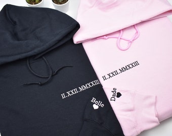 May include: Two hooded sweatshirts, one black and one light pink, with the same Roman numeral date and the words "Belle" and "Dade" embroidered on the sleeves. The black sweatshirt has light pink thread and the pink sweatshirt has black thread.
