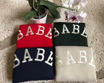 May include: Four sweatshirts with the word "BABE" embroidered in white on the front. The sweatshirts are in red, green, navy blue, and beige.