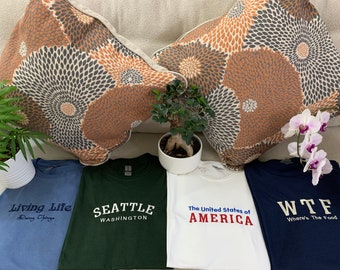 May include: Four t-shirts with text: "Living Life Doing Things", "Seattle Washington", "The United States of AMERICA", and "WTF Where's The Food". The shirts are blue, green, white, and navy. Decorative pillows and plants are in the background.
