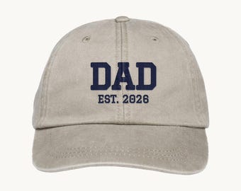 May include: A light beige baseball cap featuring the dark navy blue text DAD EST. 2026 embroidered on the front center. The cap has a curved brim, a structured crown with a button on top, and visible stitching details along the panels and brim.