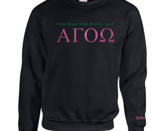 May include: A black crewneck sweatshirt featuring the text Alpha Kappa Alpha Sorority, Inc. in green, with the Greek letters ΑΓΟΩ in pink below it. The left sleeve cuff displays the pink text EST.2024. The garment has a Gildan brand label.