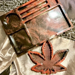 May include: A black and red resin rolling tray with a leaf-shaped compartment. The tray has a swirl pattern and a small round compartment.