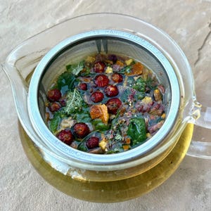 May include: A clear glass teapot with a stainless steel infuser filled with tea leaves, berries, and herbs. The tea is a light amber color. The infuser is marked "13 HARIO CHJM 70 V86" and "Made in China".