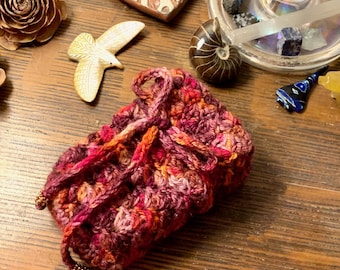 May include: A handmade crochet pouch in shades of red, orange, and purple, with a drawstring closure. The pouch is adorned with small beaded accents. The background includes decorative objects like a bird figurine and crystals.