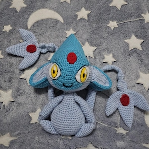 Pode incluir: Um peluche de croché azul-claro com uma cabeça pontiaguda com um remendo circular vermelho, olhos grandes amarelos com pupilas pretas e dois apêndices longos e finos nas laterais, cada um terminando numa ponta de três gumes com um círculo vermelho.