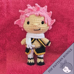 May include: Crocheted amigurumi doll with pink hair, a white scarf, and a black and gold outfit. The doll is standing on a red fuzzy surface. The text "PDF PATTERN BUNDLE" is visible in the lower right corner of the image.