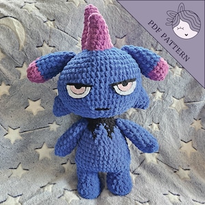 May include: A crocheted blue and purple plush toy with a grumpy expression. The toy has a horn on its head and a black collar. The toy is sitting on a gray and white blanket with stars.