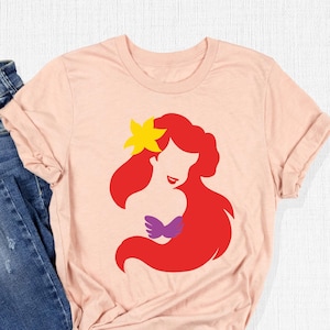 May include: A pink t-shirt with a silhouette of Ariel, the Little Mermaid, in red, purple, and yellow. The shirt is displayed on a white surface with a pair of blue jeans.