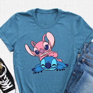 K&ouml;nnte beinhalten: Ein t&uuml;rkisfarbenes T-Shirt mit einem pinkfarbenen und blauen Stitch-Charakter-Motiv. Das Shirt liegt auf einer wei&szlig;en Oberfl&auml;che mit einem Paar schwarzer Turnschuhe und einer blauen Jeans.