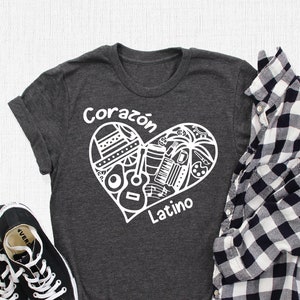 May include: A dark heather grey t-shirt with a white heart design. The heart is filled with various Latin American cultural icons, including a sombrero, a guitar, a maraca, and a palm tree. The text "Corazon Latino" is written above and below the heart.
