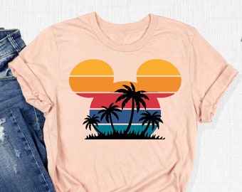 Könnte beinhalten: Ein pinkfarbenes T-Shirt mit einem Retro-Sonnenuntergangsmuster in Form von Mickey Mouse' Kopf. Palmen sind vor dem Sonnenuntergang silhouettiert. Das Shirt wird mit einer blauen Jeans kombiniert.