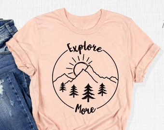 May include: A peach-colored t-shirt with a black graphic of a mountain range, sun, and trees inside a circle. The text "Explore More" is below the circle.